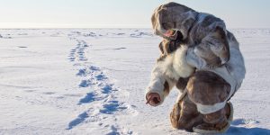 Inuit person dressed in full traditional fur outfit, tracking animal prints in the snow in Nunavut.